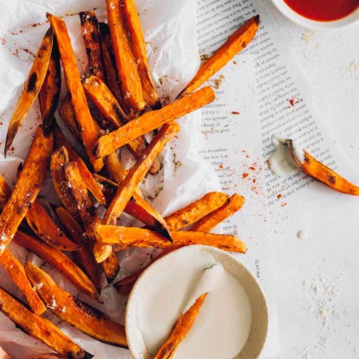 white table with some newspaper and parchment paper on which lay a bunch of crispy baked sweet potato fries next to some BBQ sauce and cashew sour cream