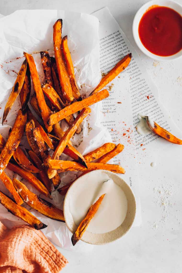 Table blanche avec un peu de papier journal et de papier sulfurisé sur laquelle sont posées un bouquet de frites de patate douce cuites au four croustillantes à côté d'un peu de sauce BBQ et de crème aigre de noix de cajou