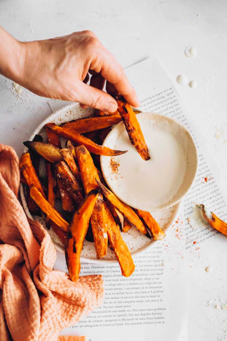 Mesa blanca con un poco de papel y un plato con patatas fritas de boniato de color naranja sin aceite, una de las cuales está siendo sumergida en crema agria vegana por una mano