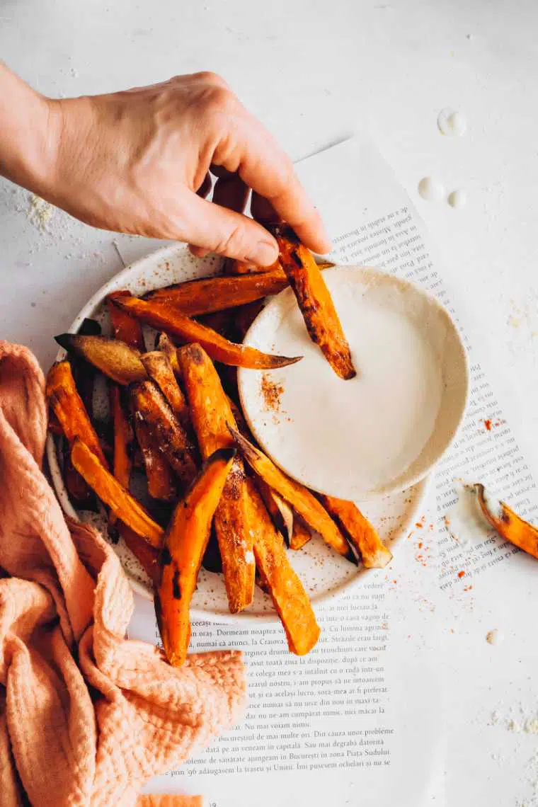 white table with some paper and a plate with oil-free orange sweet potato fries one of which is being dipped into vegan sour cream by a hand