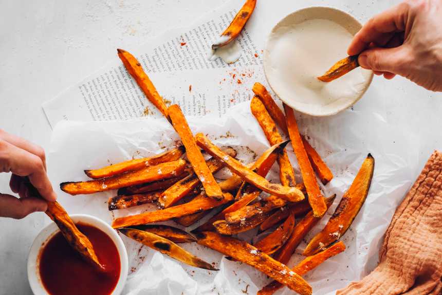 white table with parchment paper on which there is a bunch of crispy baked oil-free sweet potato fries which are being dipped in vegan BBQ sauce and cashew sour cream by two hands