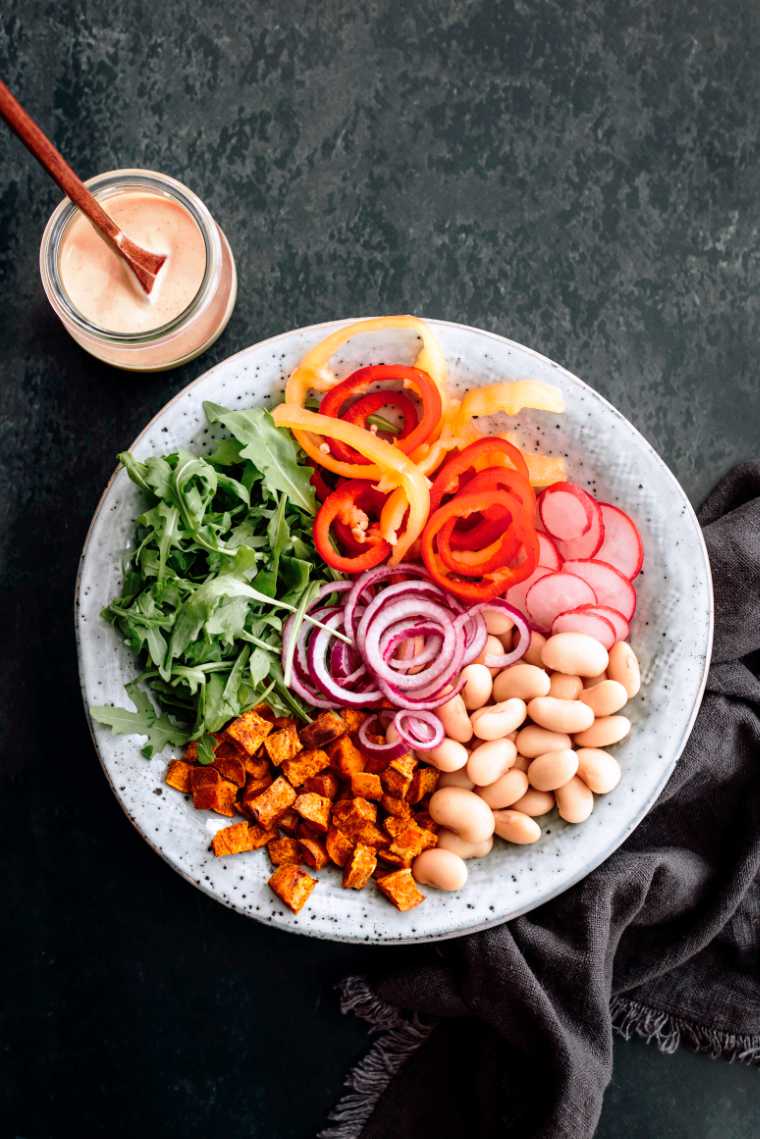 white bowl with roasted sweet potatoes, lima beans, rocket, sliced bell peppers and onion next to a vegan mustard dressing