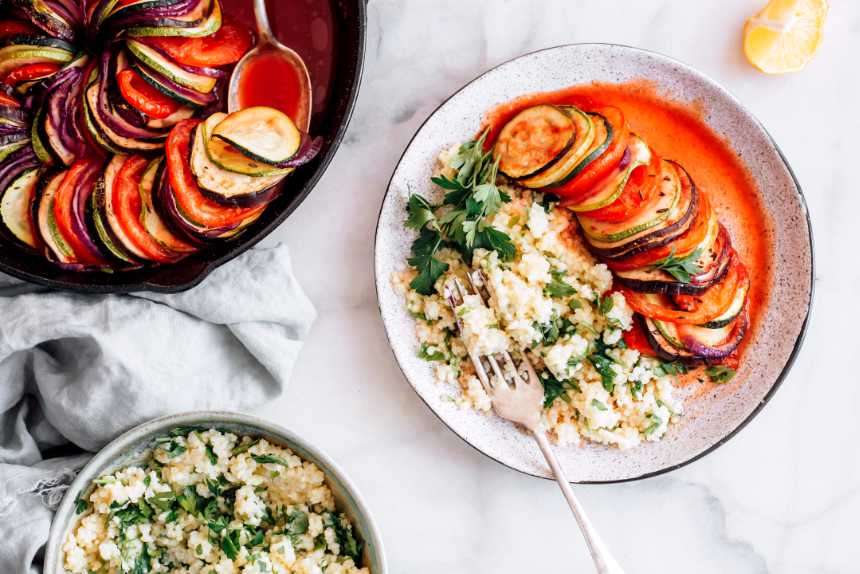 table with freshly baked vegan tian in a baking dish next to a bowl of millet and a plate with the baked ratatouille, millet, herbs and a fork