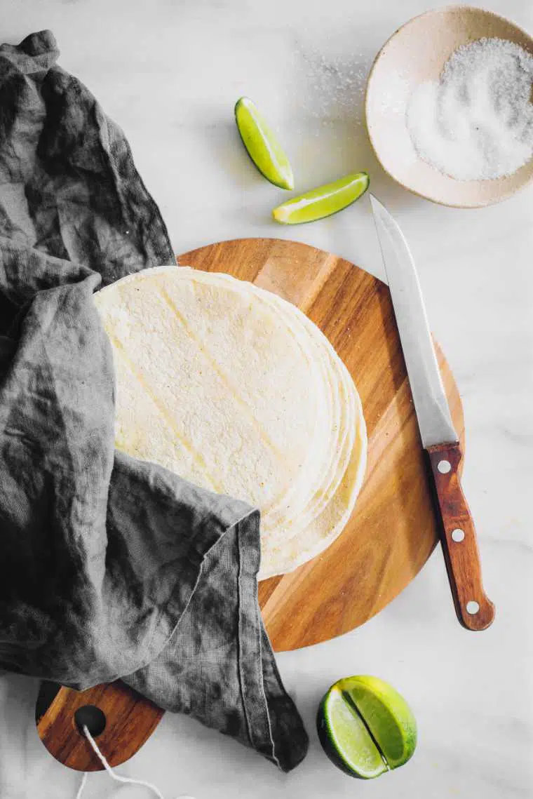 white table with a grey towel, sliced lime and a wooden cutting board with corn tortillas