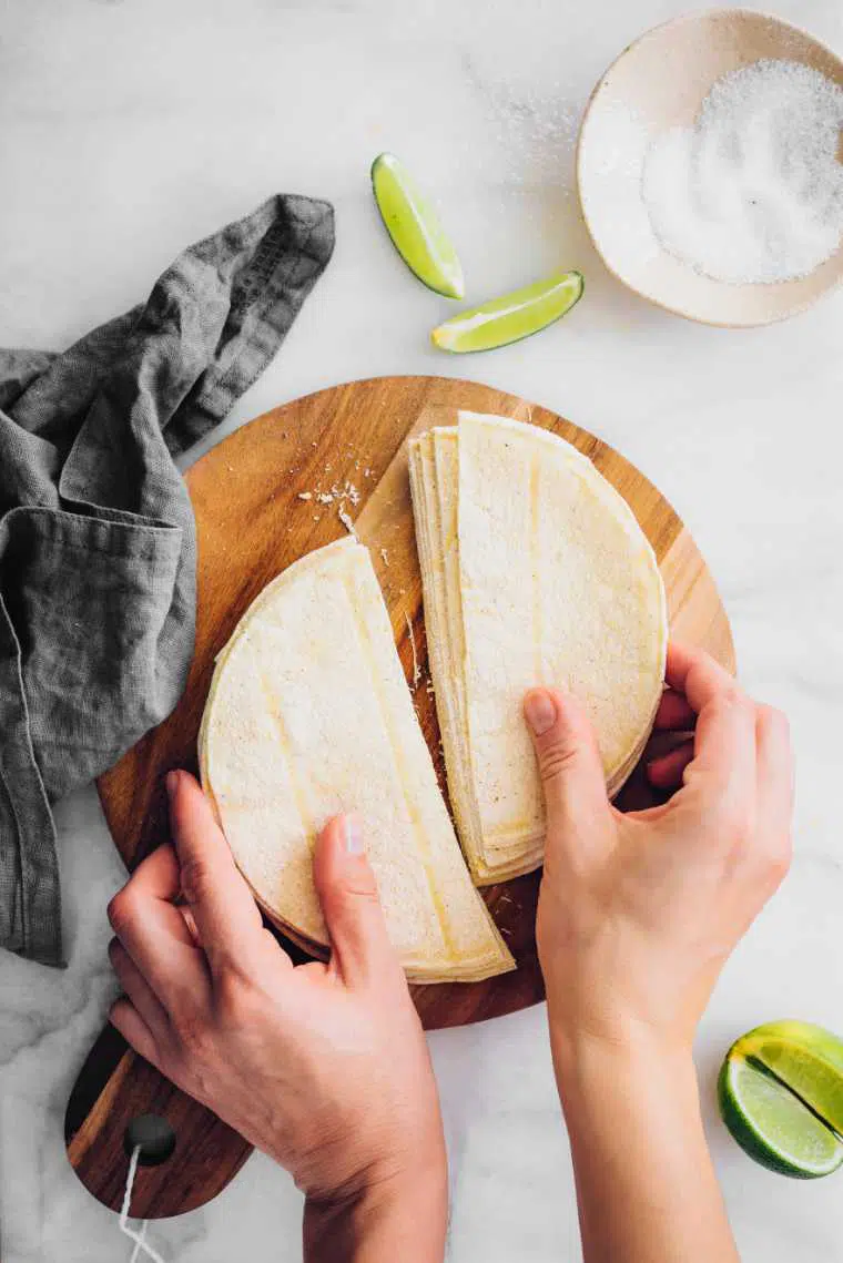 white table with wooden cutting board and halved corn tortillas held by hands