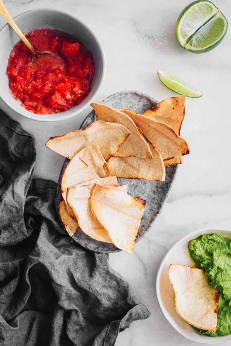freshly baked oil-free tortilla chips in a small bowl next to some guacamole and red salsa