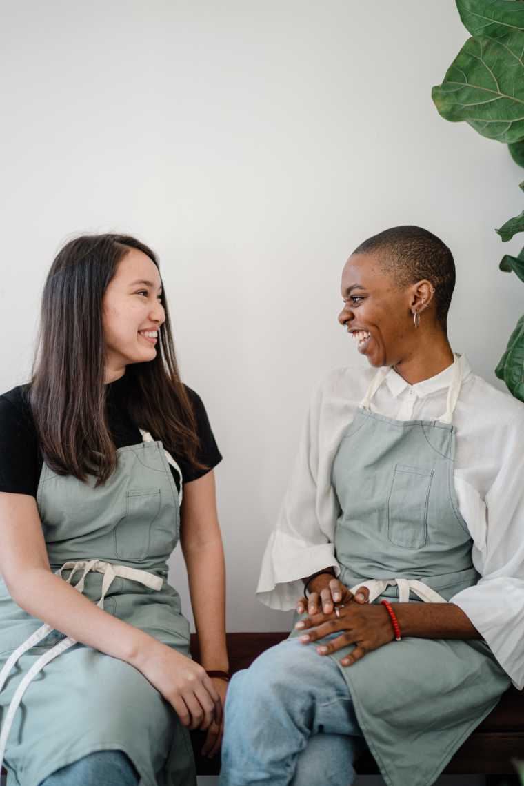two women sitting in front of a white wall and talking while smiling