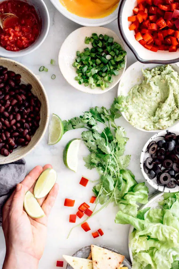 white table with bowls of vegan food like black beans, chopped bell pepper, olives, green onion, guacamole and salsa