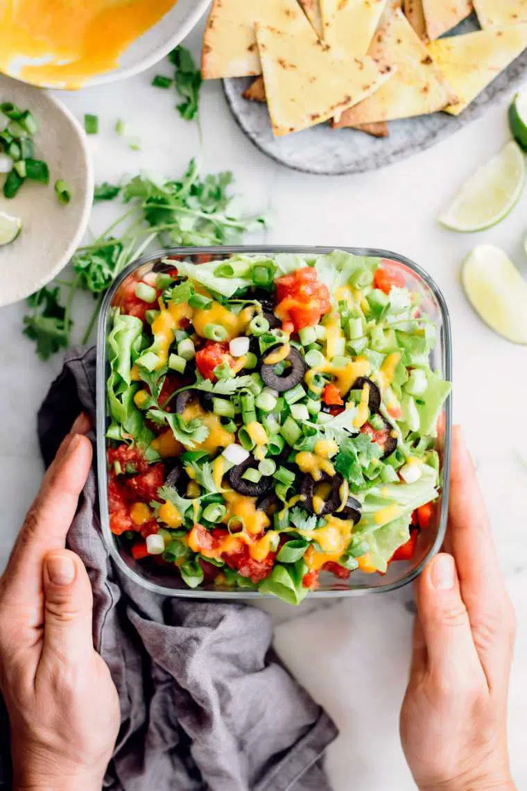 two hands holding healthy 7 layer taco dip next to oil free tortilla chips