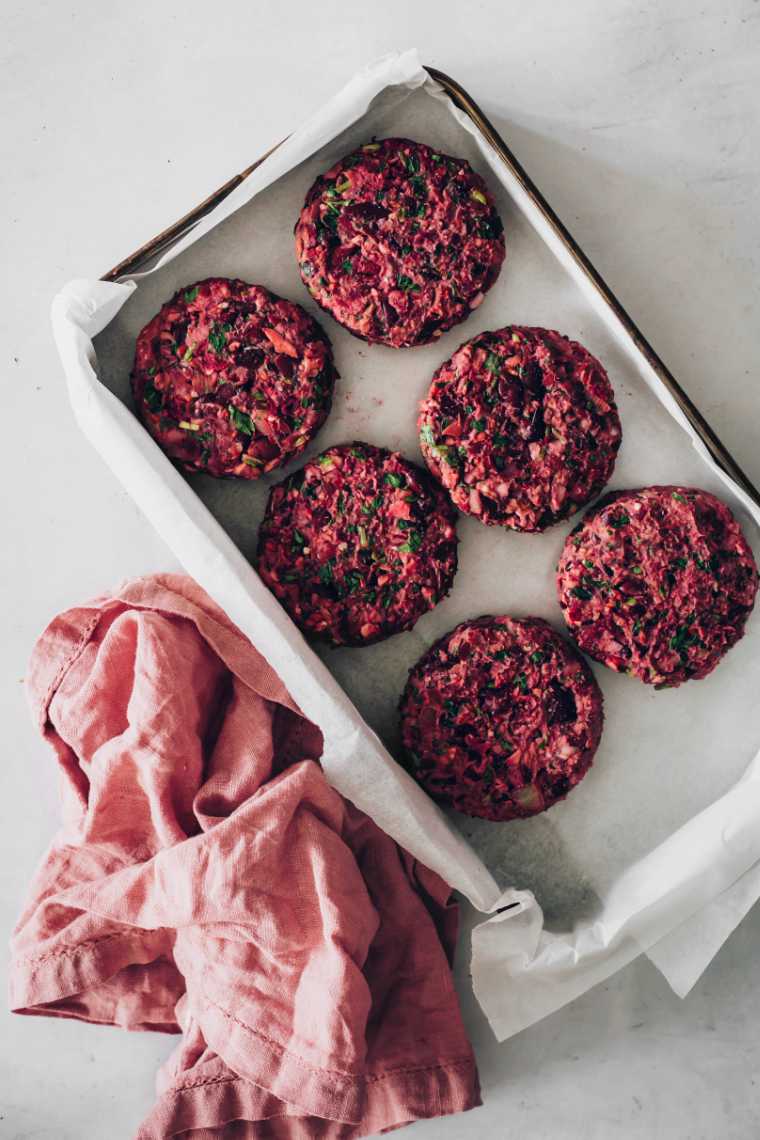 six purple-colored beetroot and kidney bean burger patties next to each other on some parchment paper in a baking dish