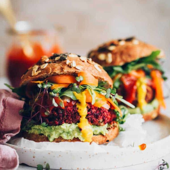 two large and colorfully stuffed beet burger patties in whole wheat buns with avocado, mustard, BBQ sauce, greens and carrot on a plate