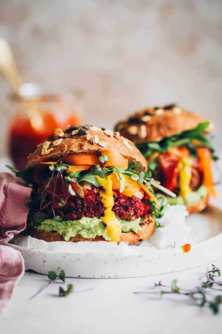 two large and colorfully stuffed beet burger patties in whole wheat buns with avocado, mustard, BBQ sauce, greens and carrot on a plate