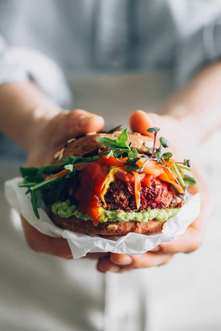 woman in blue shirt holding an assembled vegan burger with avocado, mustard and BBQ sauce in her hands