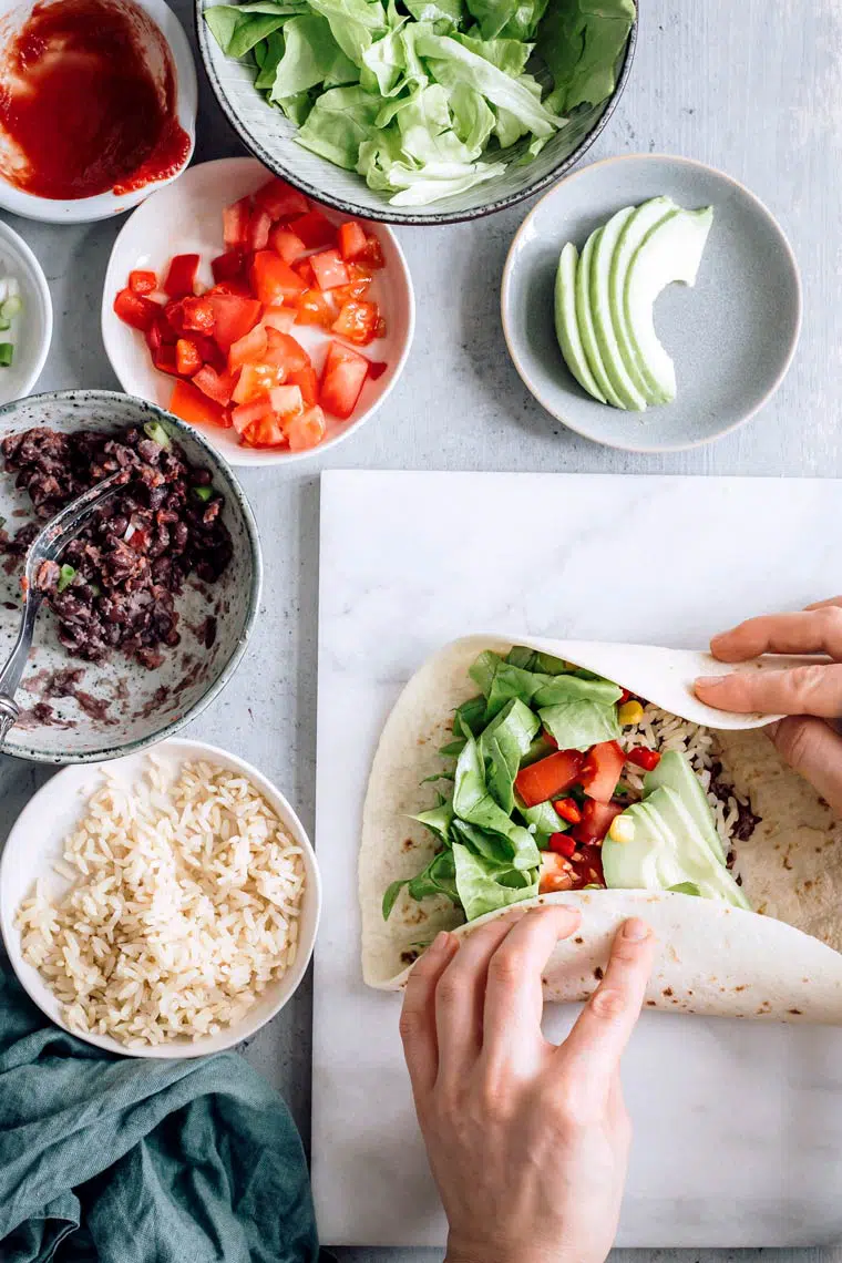 Woman rolling bean burrito on cutting board next to small bowls with vegetables