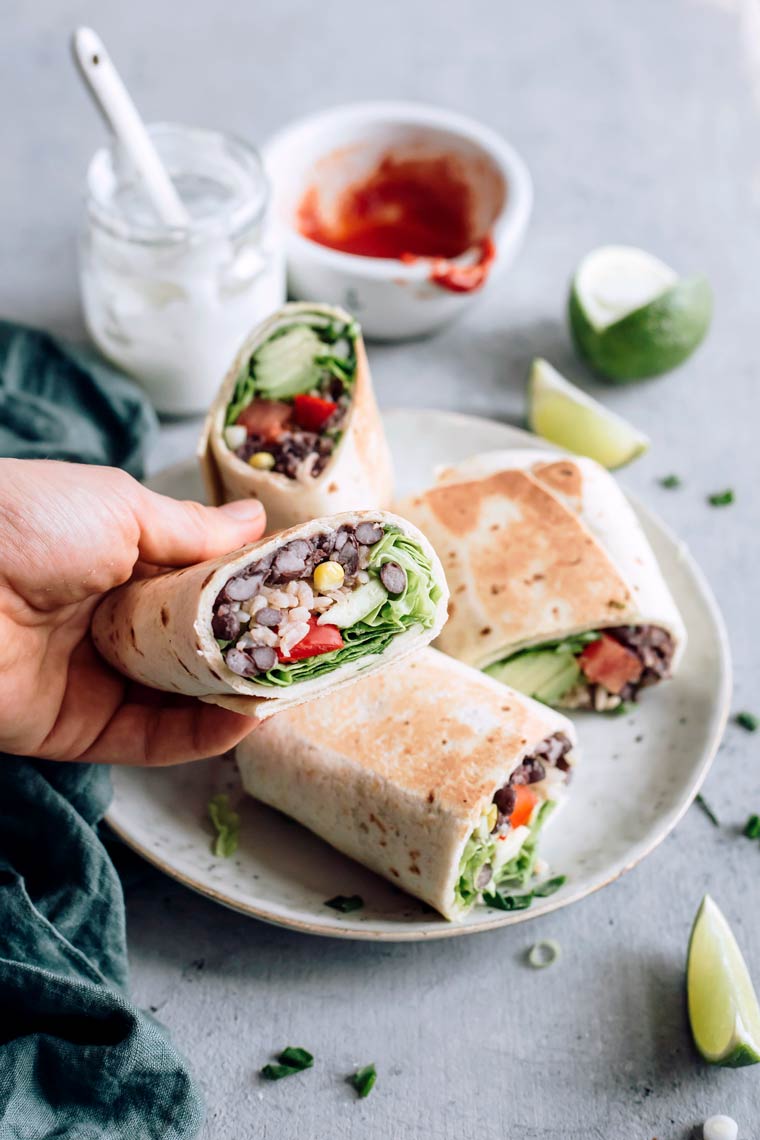 Woman holding halved bean burrito in her hand over a plate of two more burritos