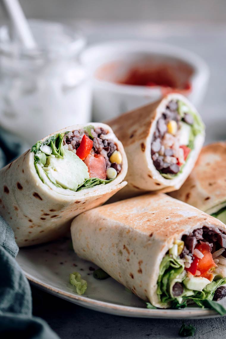 Close up of four bean burritos filled with vegetables on a plate with yogurt and tomato paste in the background