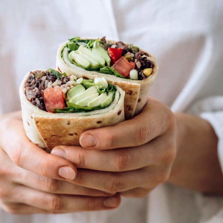 woman with white shirt holding two bean burritos filled with avocado and tomato