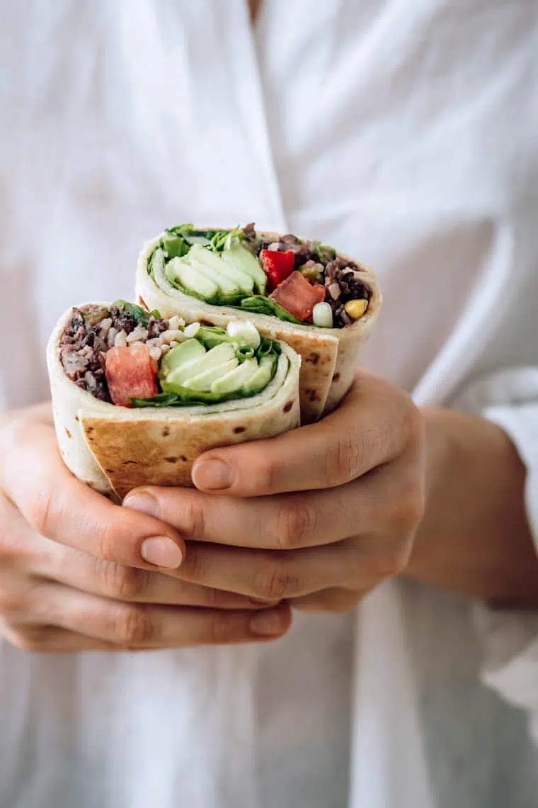 woman with white shirt holding two bean burritos filled with avocado and tomato