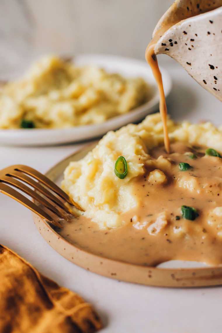 two plates with mashed potatoes one of which is being topped with vegan brown gravy