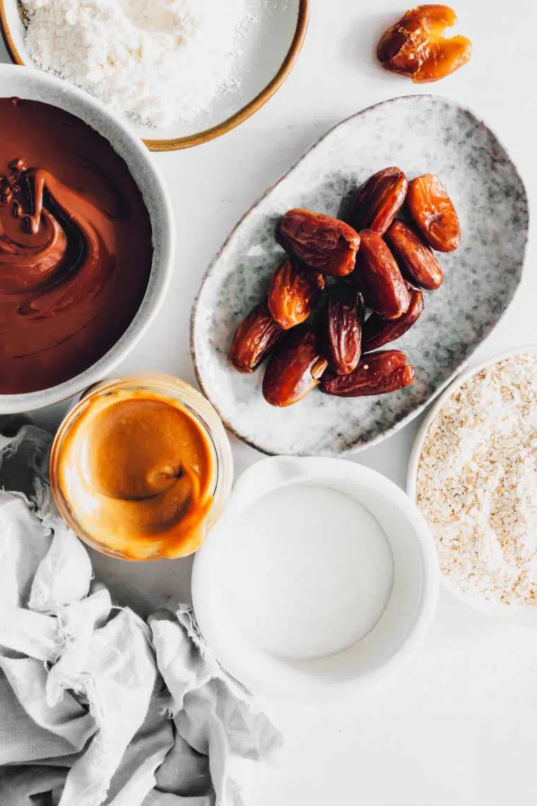 white table with different sized bowls containing ground oats, dates, almond flour, melted vegan chocolate and date caramel