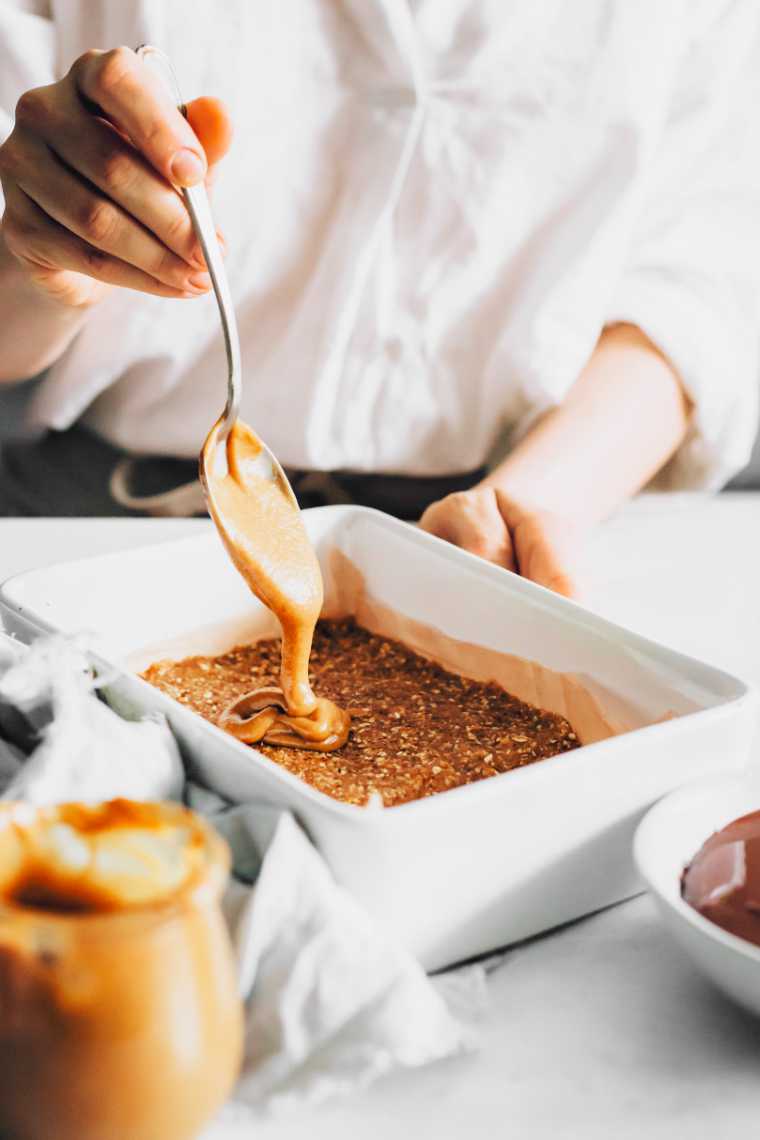 woman in white shirt standing by a table and pouring date caramel from a spoon onto almond cookie layer in a baking dish