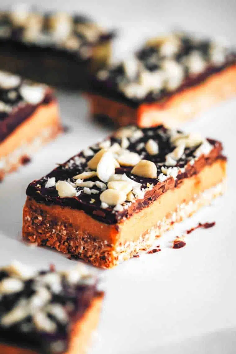 five no-bake vegan caramel slices with chocolate and almond topping on a white table
