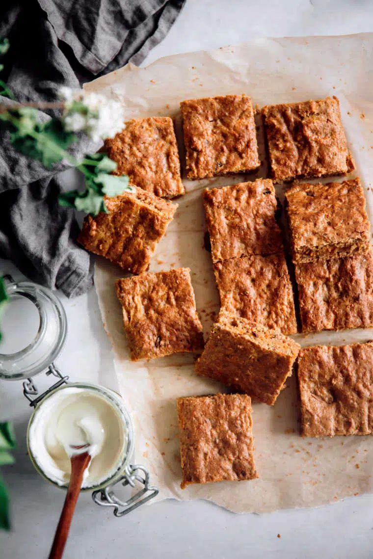 12 squares of freshly baked plant based carrot cake on parchment paper next to some cashew frosting