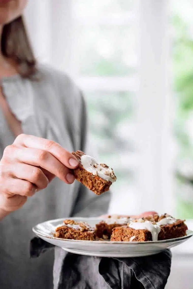 woman standing by a window with a place of plant-based carrot cake pieces and taking one to eat