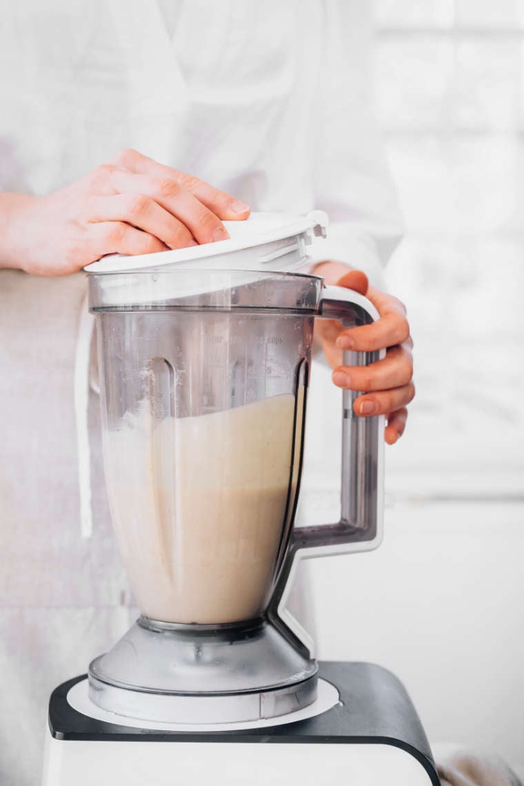 woman in white apron standing behind a blender jar with freshly made cashew mayo