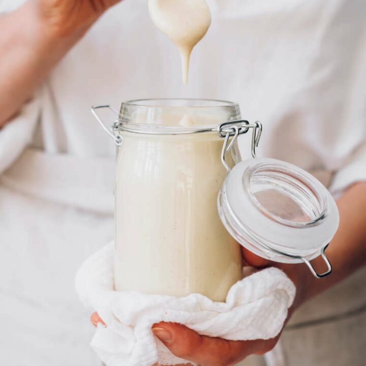 woman in white shirt holding a glass jar containing creamy cashew mayo and dipping a spoon into it