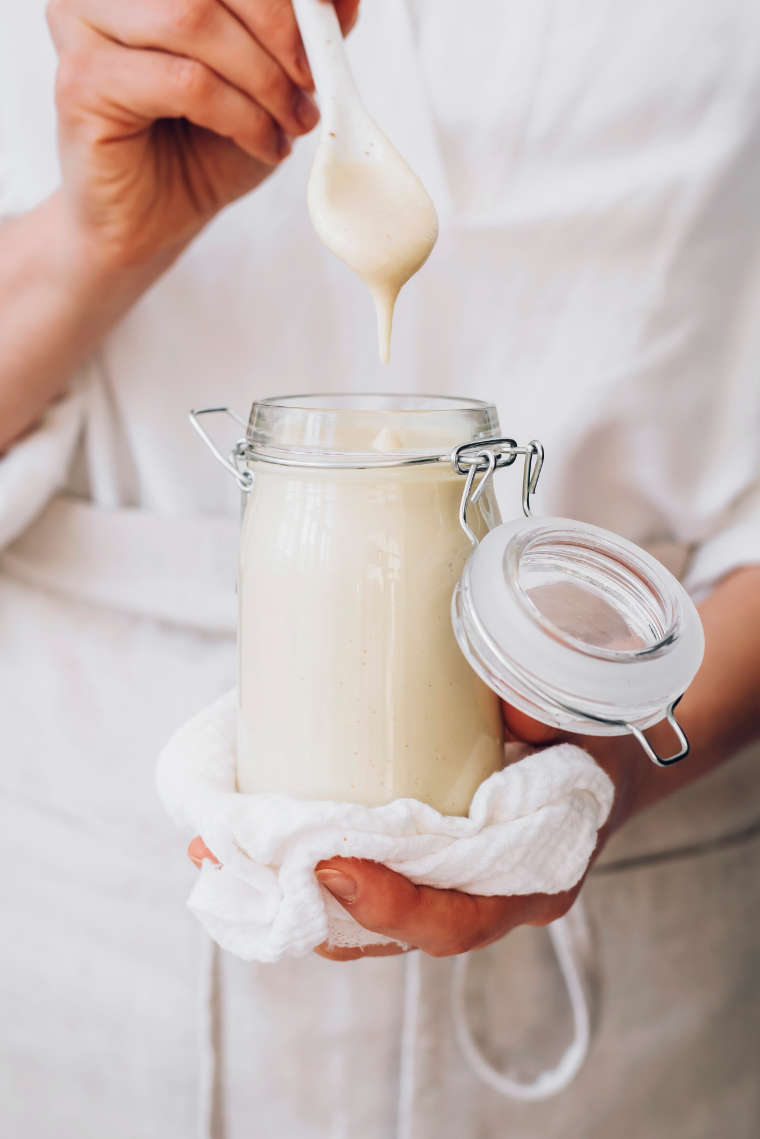 woman in white shirt holding a glass jar containing creamy cashew mayo and dipping a spoon into it