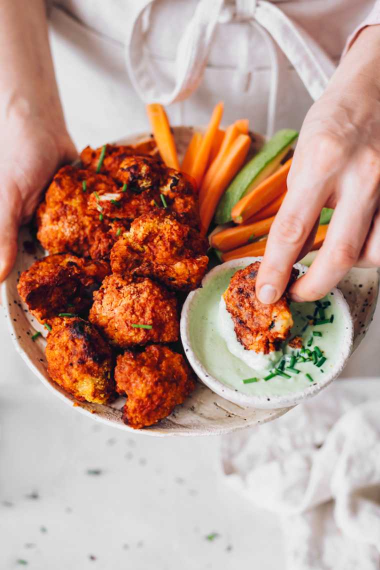 woman in apron holding a white plate with buffalo cauliflower while dipping one of the florets into a vegan ranch