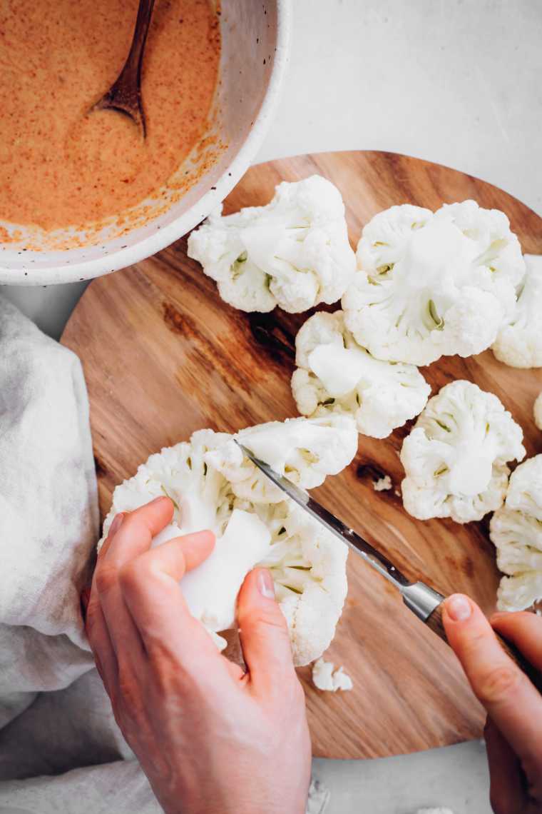 wooden cutting board on a white surface and two hands cutting cauliflower in florets with a knife