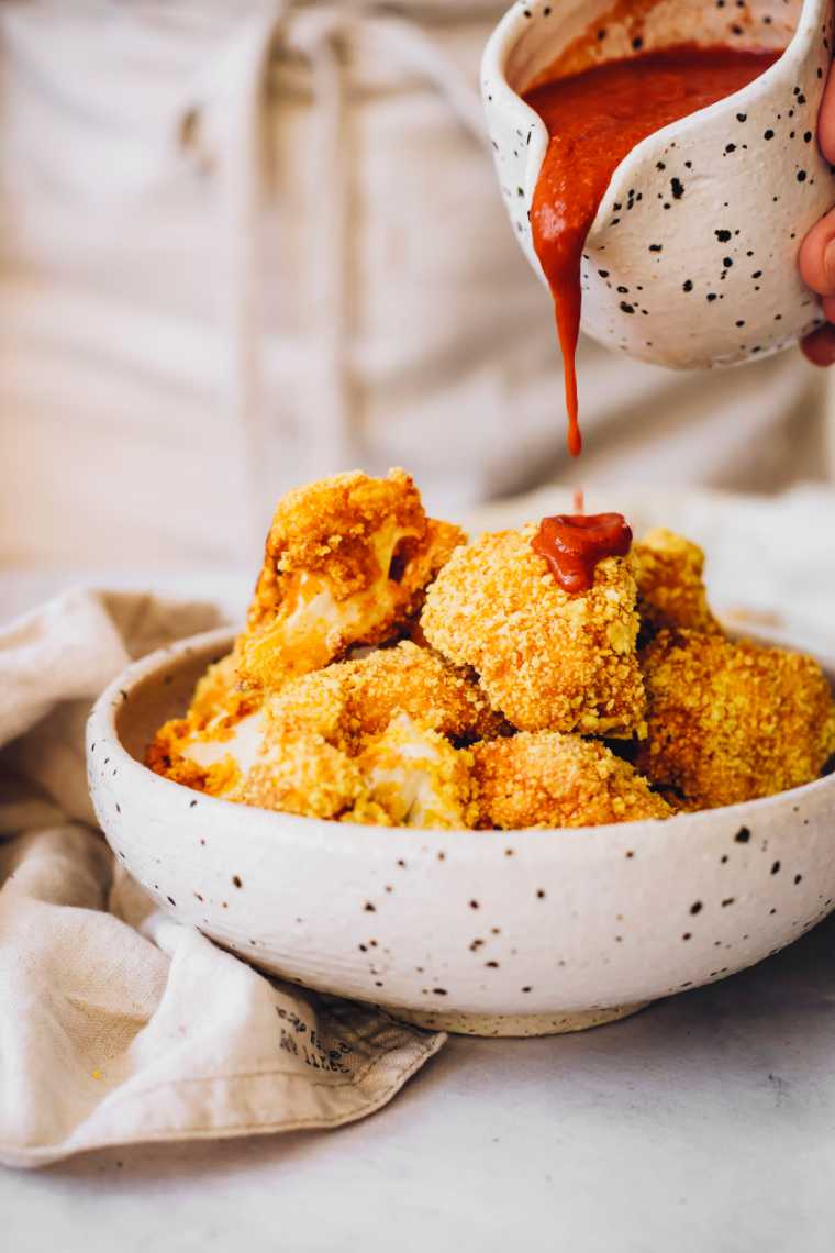 white speckled bowl with baked cauliflower bites that are being drizzled with homemade vegan buffalo sauce