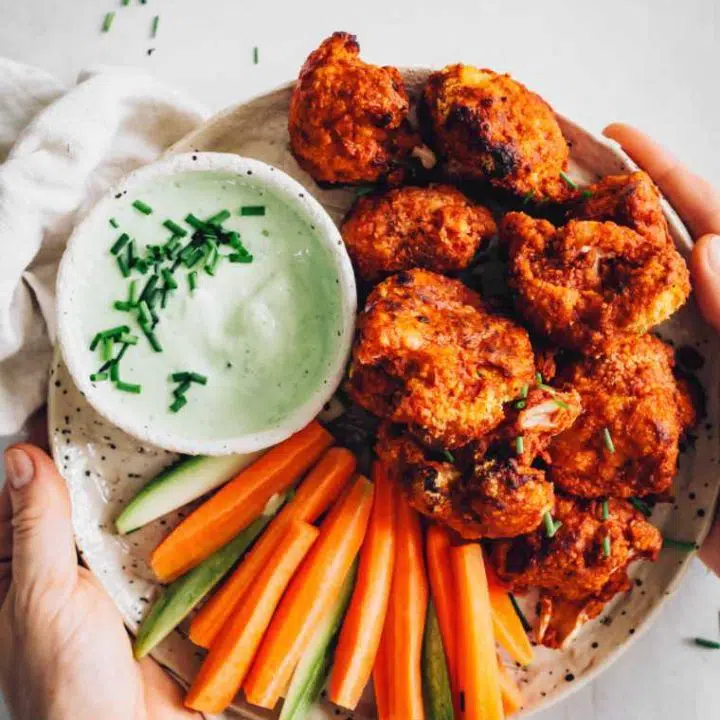 two hands holding a white plate with crispy baked vegan chicken wings next to carrot and zucchini sticks as well as green vegan ranch dip