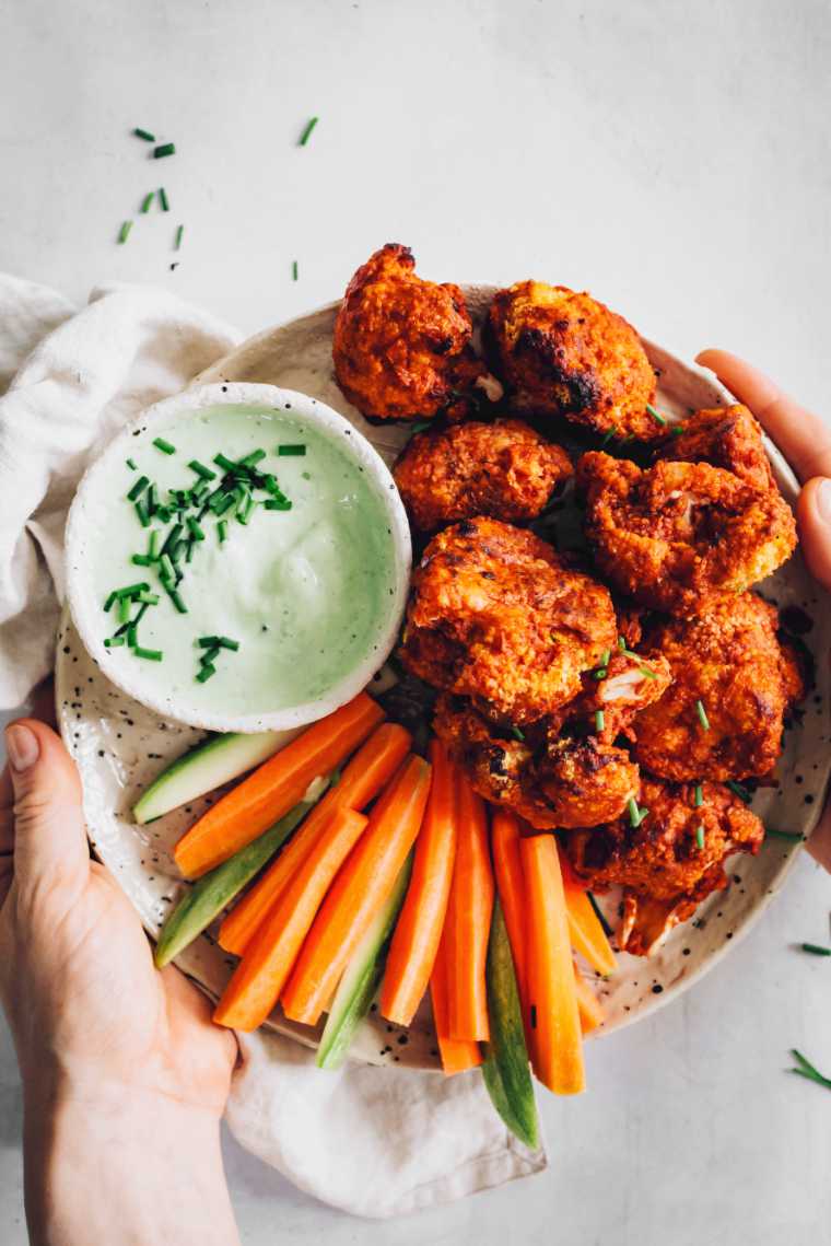 two hands holding a white plate with crispy baked vegan chicken wings next to carrot and zucchini sticks as well as green vegan ranch dip