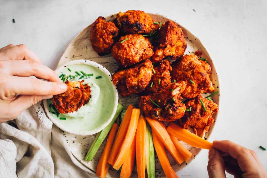 plate of crispy baked cauliflower buffalo wings next to some carrot and zucchini sticks as well as vegan ranch dip
