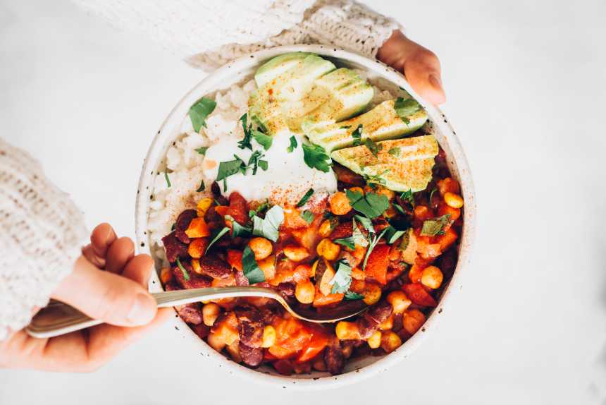 Top view of woman holding bowl filled with rice, bean chili, sliced avocado, vegan yogurt and sprinkled herbs