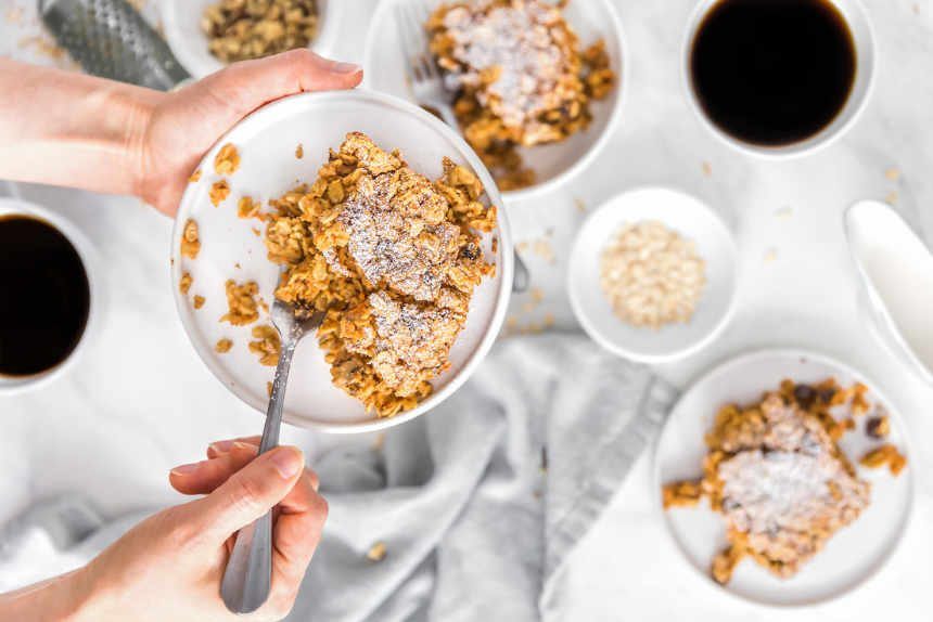 woman holding white plate with a vegan carrot cake oatmeal Christmas breakfast bake over a table with more oatmeal bake and two coffees