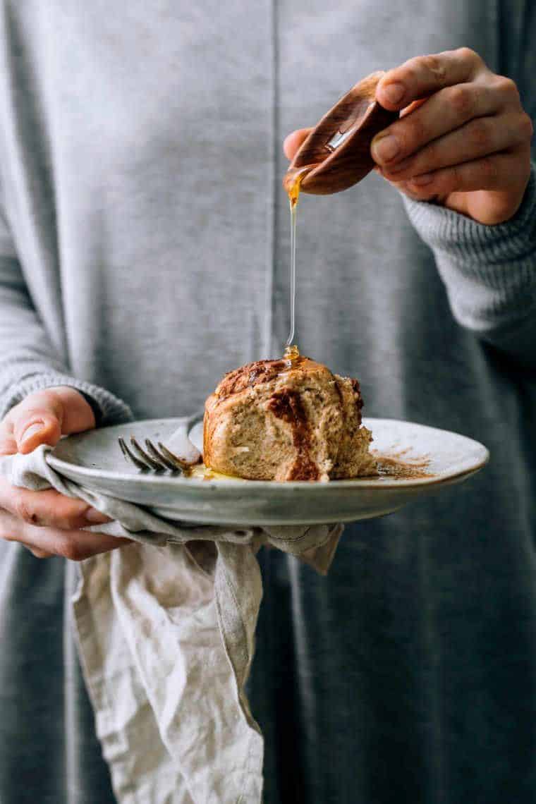 woman in grey sweater holding a white plate with a vegan cinnamon roll and pouring maple syrup over it
