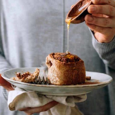woman in grey sweater holding a white plate with a vegan whole wheat cinnamon roll and pouring maple syrup over it