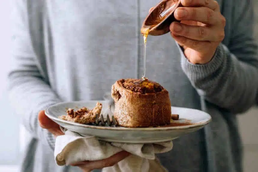 woman in grey sweater holding a white plate with a vegan whole wheat cinnamon roll and pouring maple syrup over it