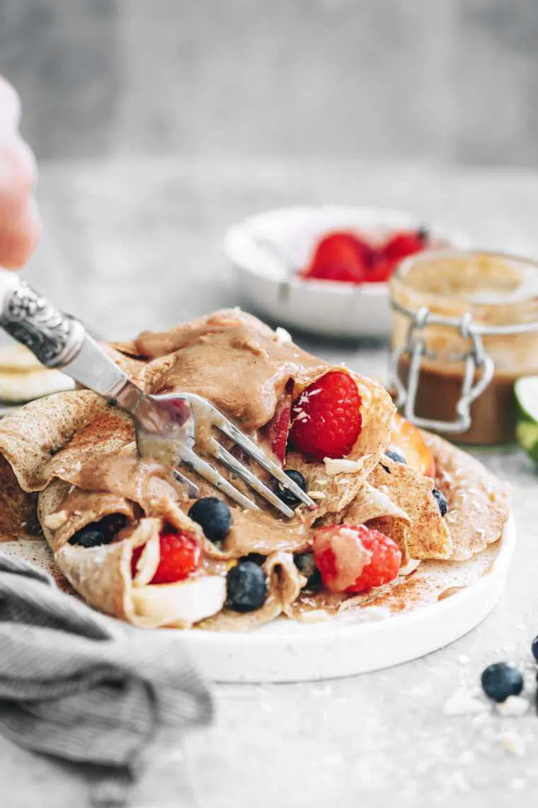 white plate with fruit stuffed almond milk crepes in which a hand cuts with a fork