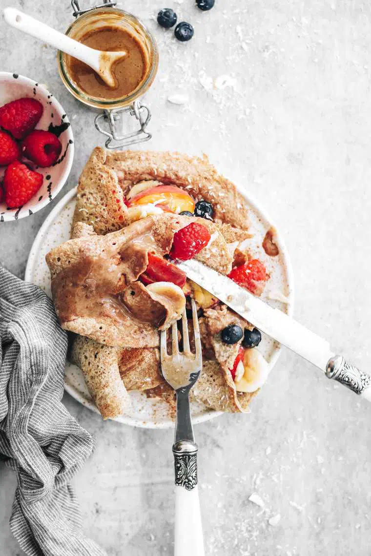 top view of a white plate with whole grain vegan crepes and fruit topped with some cutlery