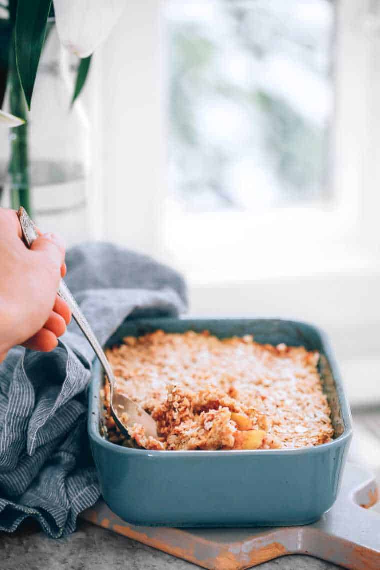 blue baking dish containing sugar-free apple crumble on a counter top and a hand taking some of it out with a spoon