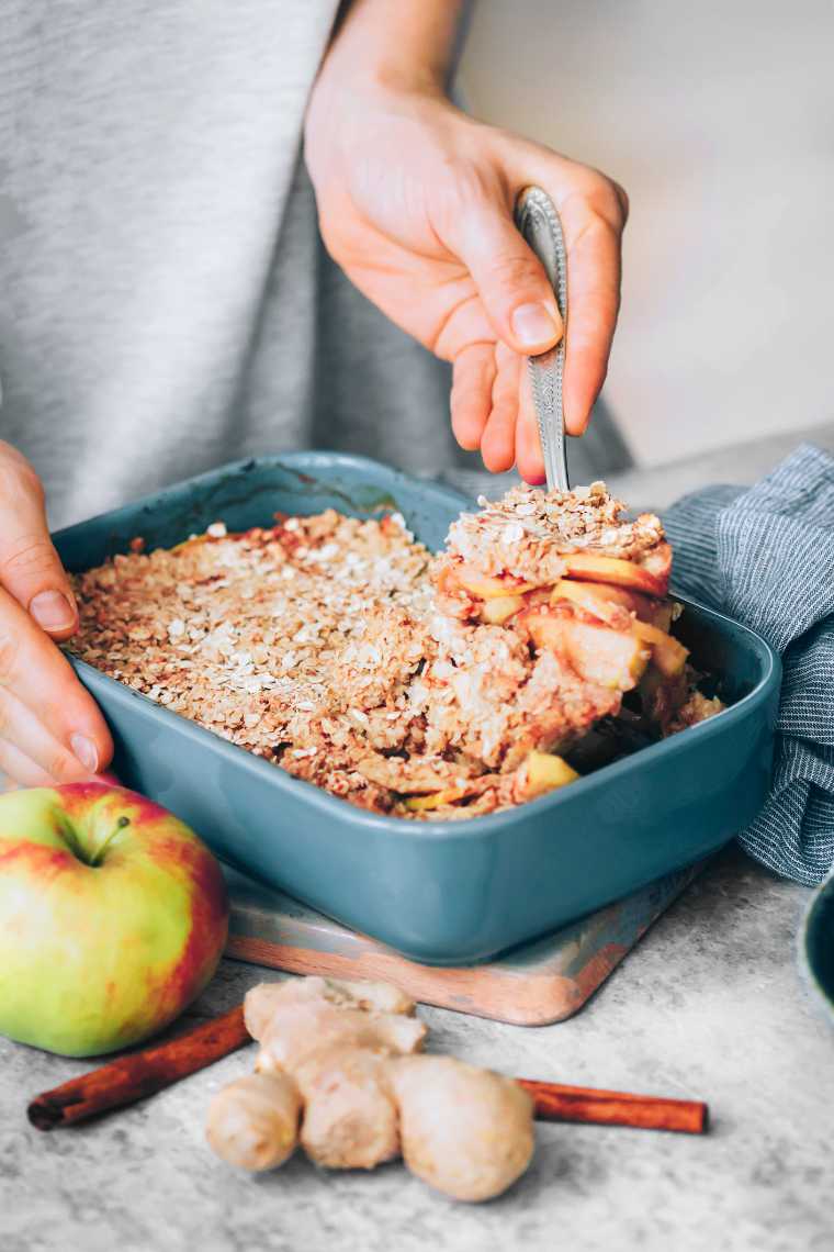 woman standing by a baking dish with sugar-free vegan apple crumble and serving it with a spoon