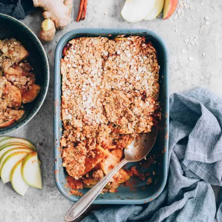 grey table with a towel, apple slices, cinnamon, ginger and a baking dish with baked vegan crumble