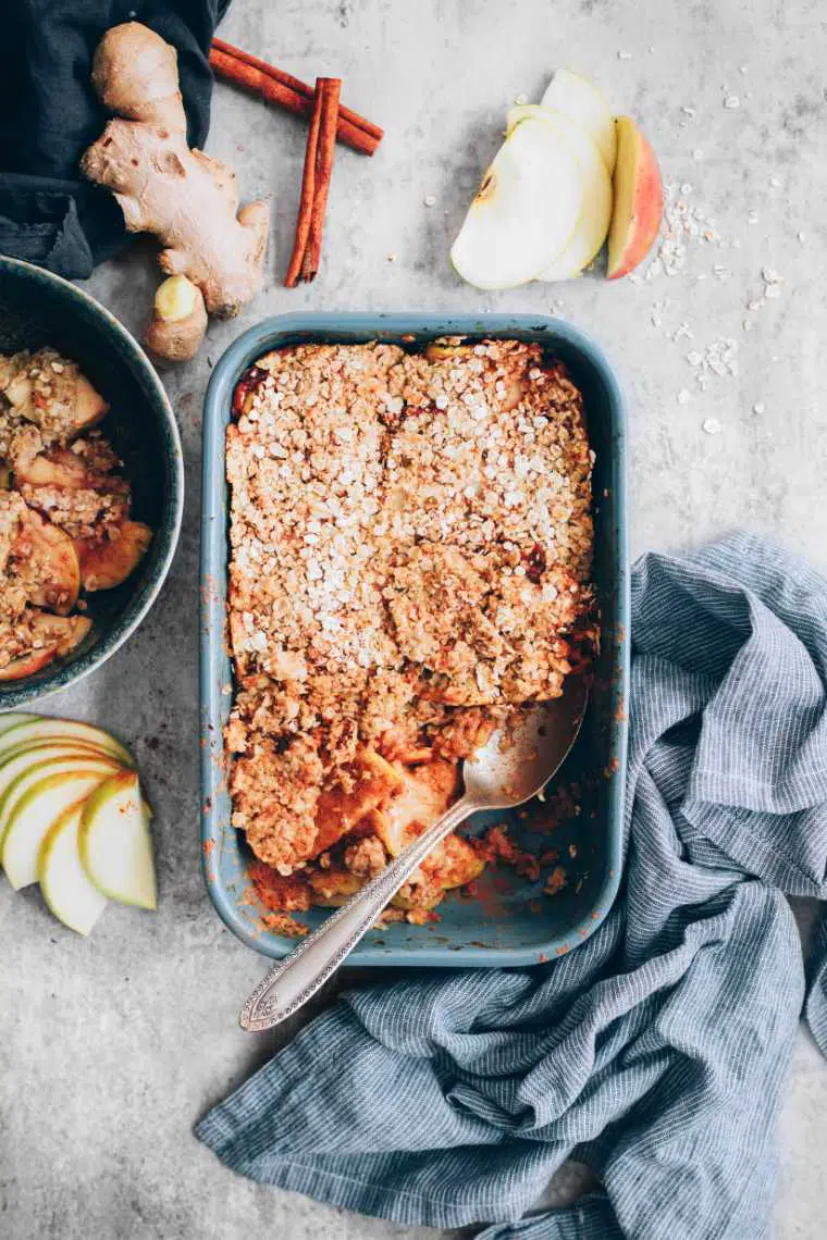 grey table with a towel, apple slices, cinnamon, ginger and a baking dish with baked vegan crumble
