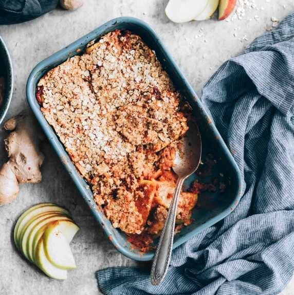 healthy vegan apple crumble in a baking dish next to apple slices, ginger and a towel