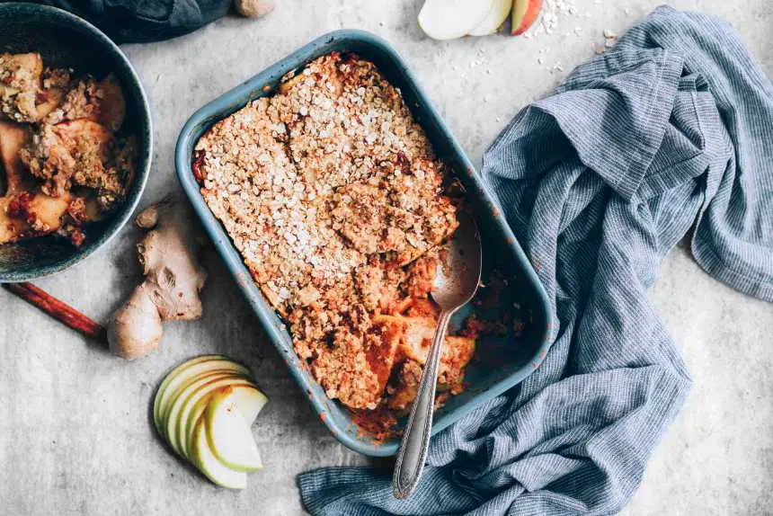 healthy vegan apple crumble in a baking dish next to apple slices, ginger and a towel